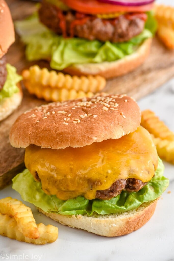 Hamburger topped with cheese on a bun with lettuce. French fries and hamburgers sitting beside and behind