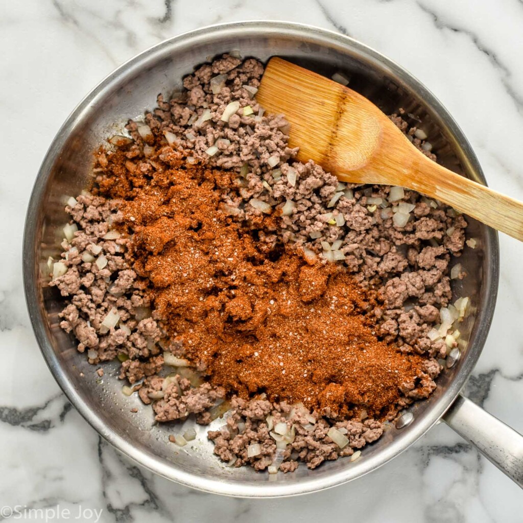 overhead of skillet of cooked ground beef with diced onions and taco seasoning to make Walking Tacos, wooden spoon for stirring.