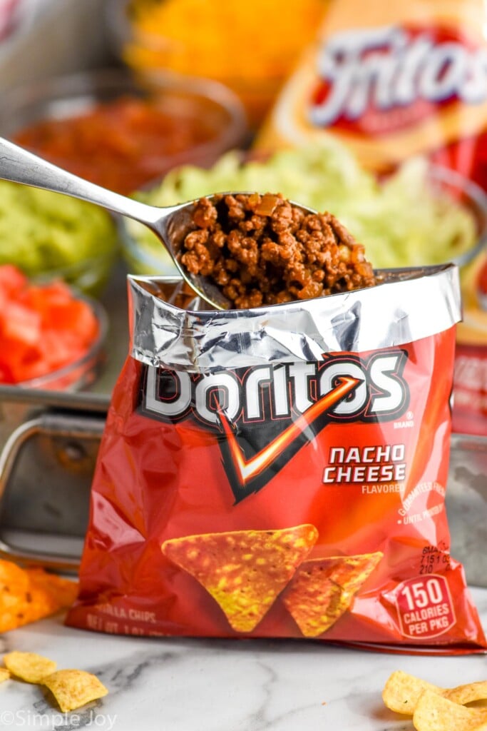 bag of doritos with spoon of taco meat going in to make Walking Tacos. Walking Tacos toppings sitting in background