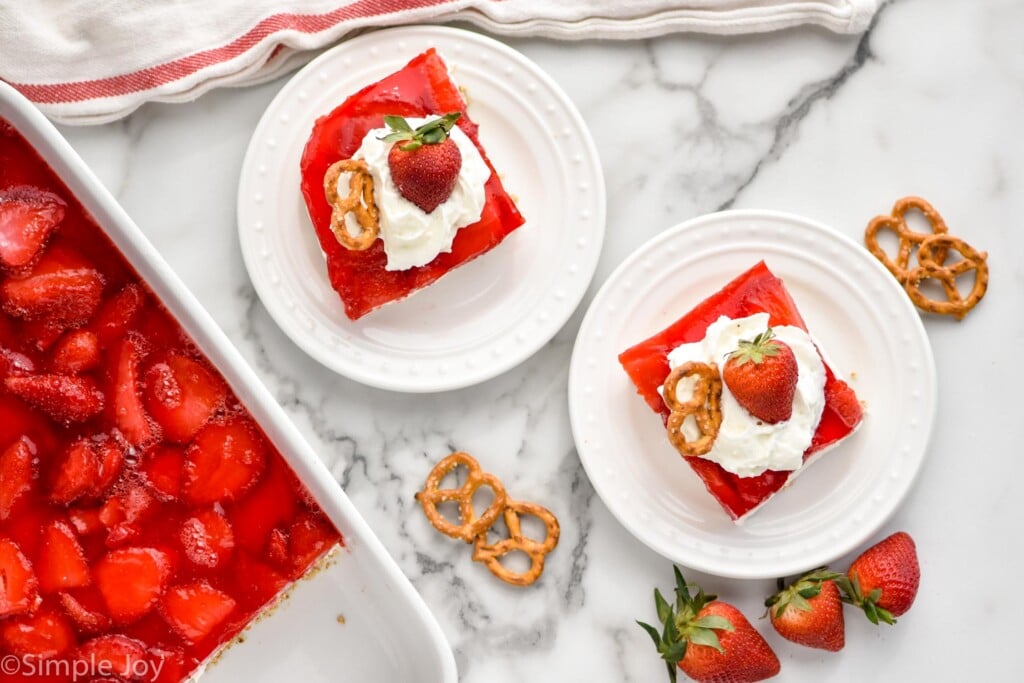 overhead of two plates of Strawberry Pretzel Salad topped with whipped cream, a strawberry, and a pretzel. Pan of Strawberry Pretzel Salad, pretzels, and fresh strawberries sitting beside