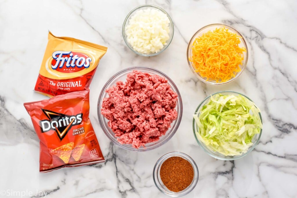 overhead of bags of doritos and fritos and bowls of ground beef, taco seasoning, shredded cheese, diced onions, and shredded lettuce to make Walking Tacos