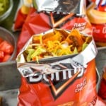 bag of doritos of walking tacos topped with shredded cheese, shredded lettuce, and taco meat. Toppings and bags of walking tacos sitting behind - 13