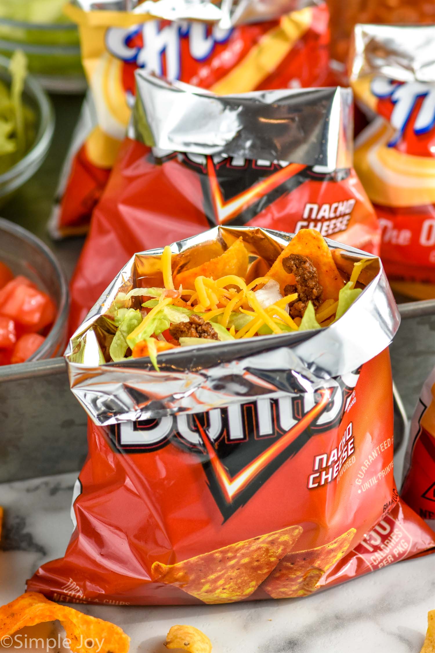 bag of doritos of walking tacos topped with shredded cheese, shredded lettuce, and taco meat. Toppings and bags of walking tacos sitting behind