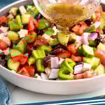Pinterest graphic for Greek Salad recipe. Image shows person's hand pouring glass cup of dressing over chopped vegetables for Greek Salad recipe. Text says, "Greek Salad simplejoy.com."