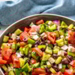 Pinterest graphic for Greek Salad recipe. Text says, "the best Greek Salad recipe simplejoy.com." Image shows a bowl of Greek Salad garnished with dressing and seasonings with serving spoons beside.