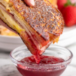 Pinterest graphic for Monte Cristo Sandwich recipe. Image shows person's hand dipping half of Monte Cristo Sandwich into a dish of sauce. Text says, "super easy Monte Cristo Sandwich simplejoy.com."