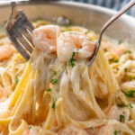 Pinterest graphic for Shrimp Alfredo recipe. Text says, "the best Shrimp Alfredo simplejoy.com." Image shows two forks lifting Shrimp Alfredo garnished with parsley flakes out of skillet.