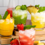 four flavors of Agua Fresca drinks served in glasses with fruit and mint leaves as garnish. More fruit and mint leaves on the counter for garnish. - 12