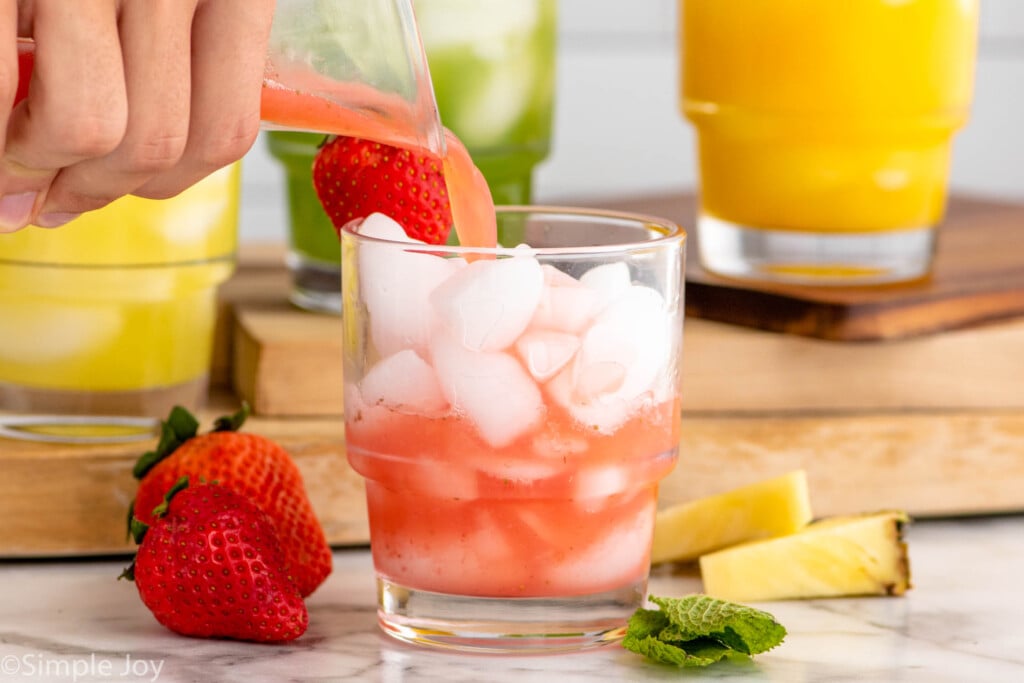 Side view of pitcher of Agua Fresca being poured into glass of ice. Strawberries, pineapple chunks, and mint leaves beside for garnish.