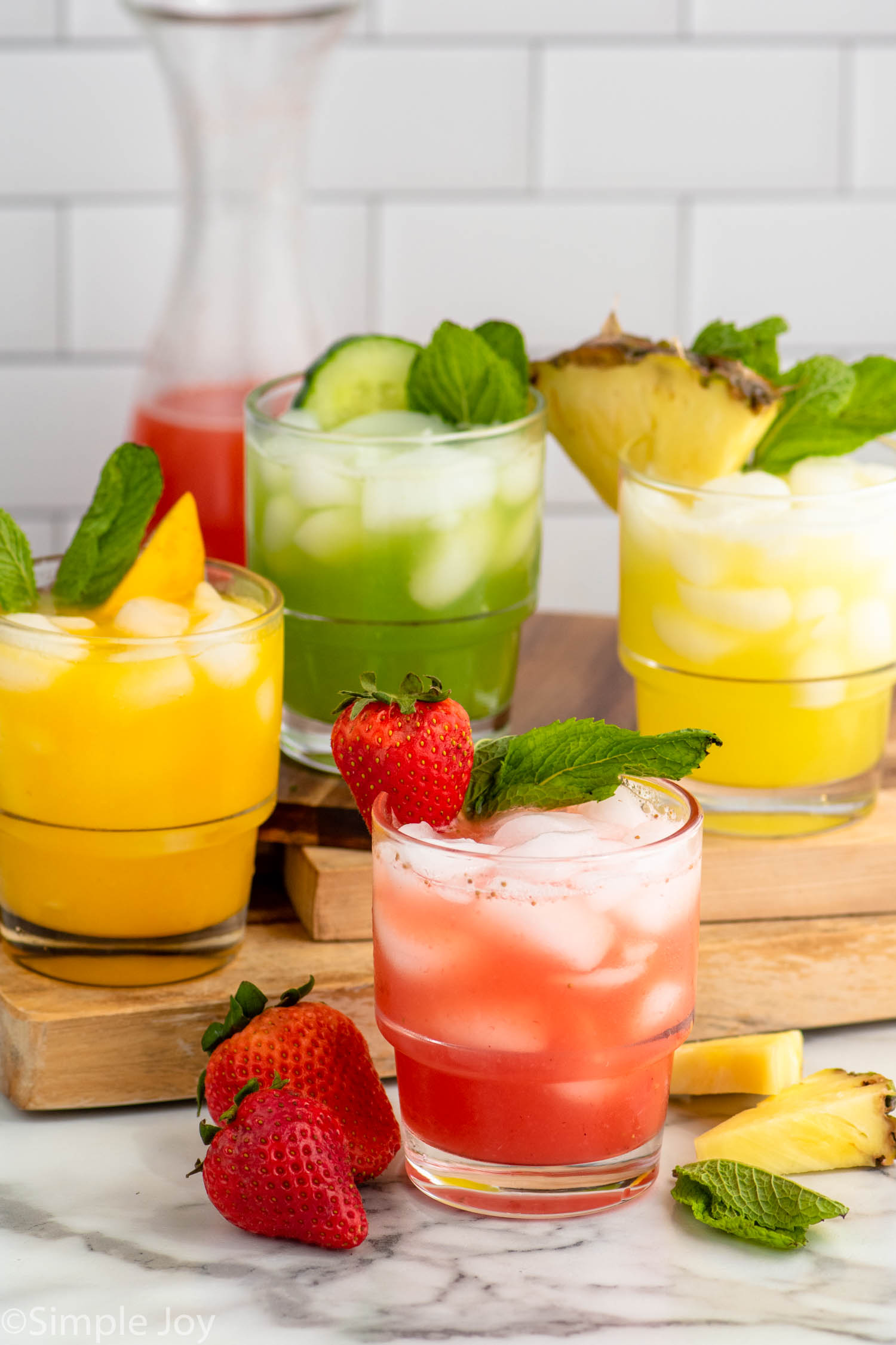 four flavors of Agua Fresca drinks served in glasses with fruit and mint leaves as garnish. More fruit and mint leaves on the counter for garnish.