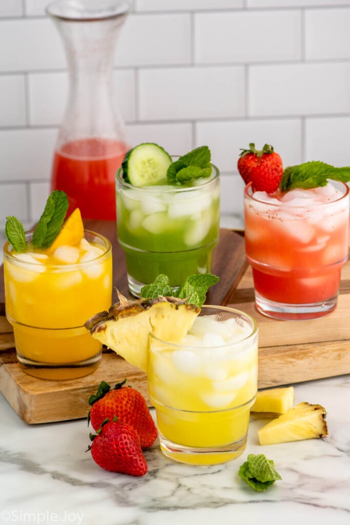 four flavors of Agua Fresca drinks served in glasses with fruit and mint leaves as garnish. More fruit and mint leaves on the counter for garnish.
