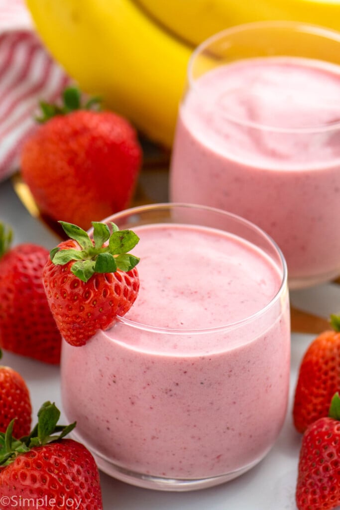 Strawberry Banana Smoothie served in glasses with strawberry on rim as garnish. Strawberries and bananas beside.