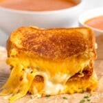 Side view of a grilled cheese sandwich cut in half with the halves stacked on top of each other. Bowls of tomato soup in the background. - 10