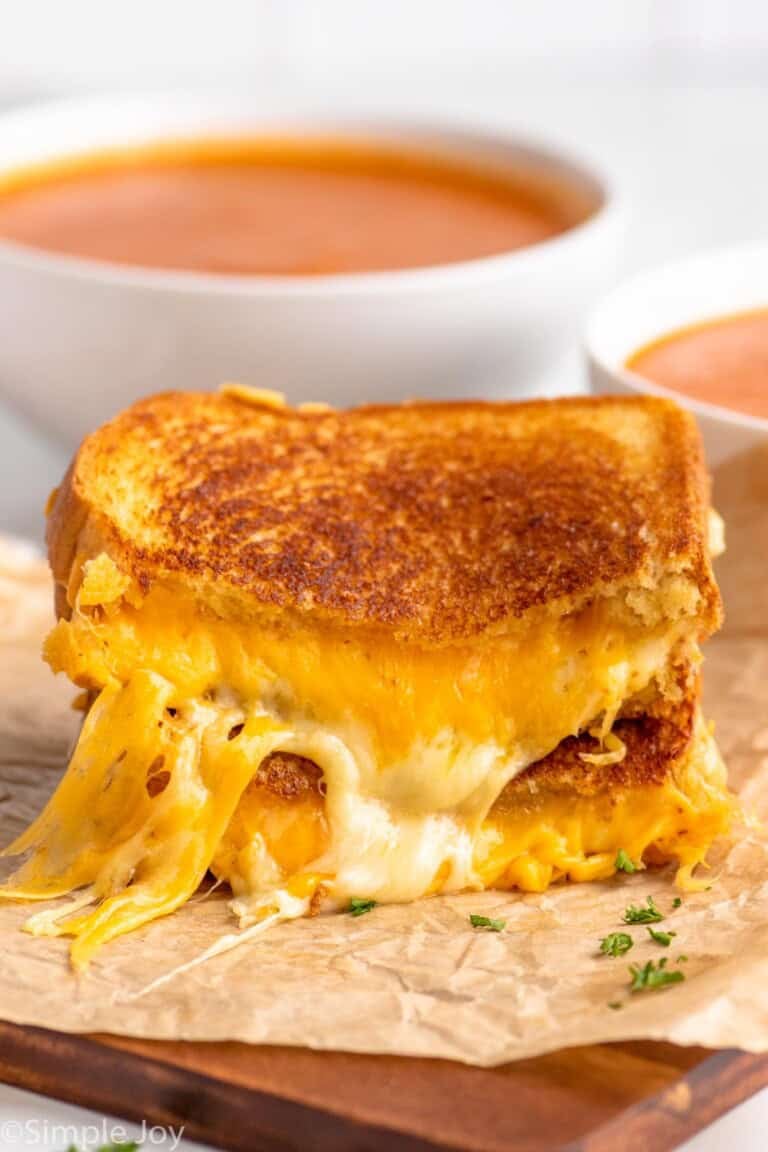 Side view of a grilled cheese sandwich cut in half with the halves stacked on top of each other. Bowls of tomato soup in the background.
