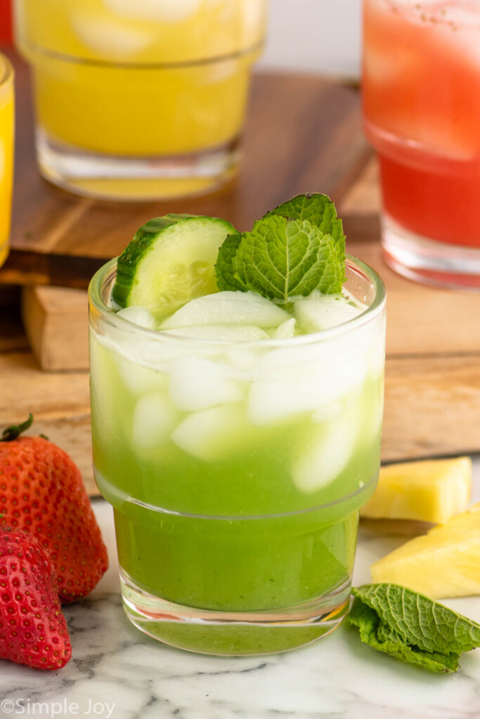 Cucumber Agua Fresca garnished with cucumber slice and mint leaves. Strawberries, pineapple chunks, and mint leaves beside for garnish.