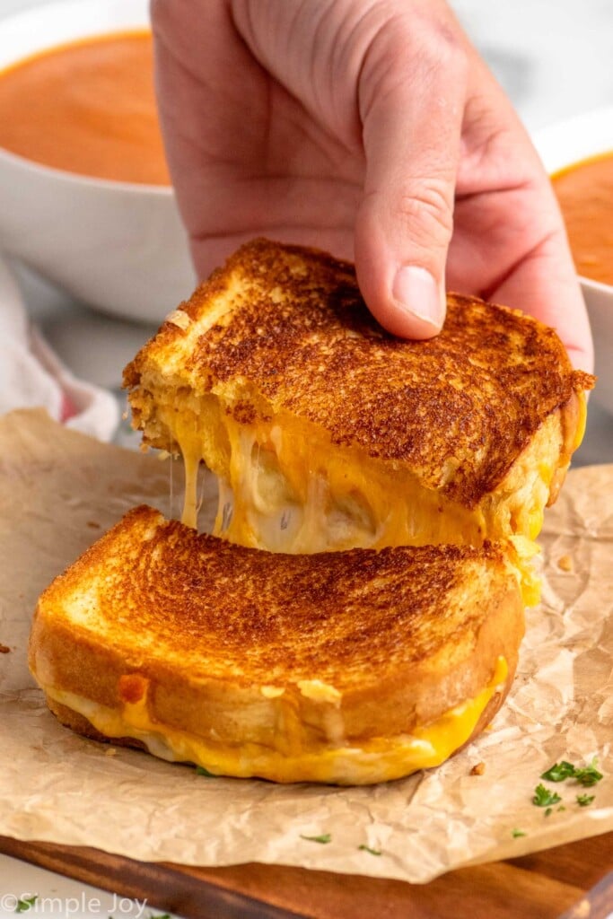 Person's hand pulling apart two halves of a grilled cheese sandwich with bowls of tomato soup beside