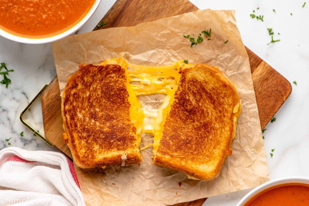 Overhead view of grilled cheese sandwich cut in half and pulled apart with cheese between. Bowls of tomato soup beside