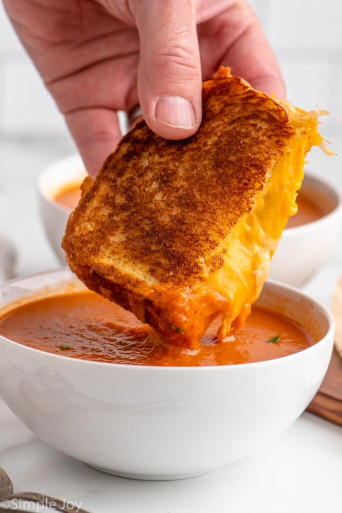 Person's hand dipping half of a grilled cheese sandwich into a bowl of tomato soup