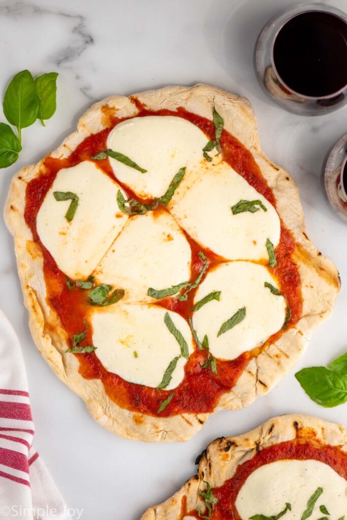 Overhead view of Grilled Pizza on counter topped with cheese and shredded basil with glass of red wine beside.