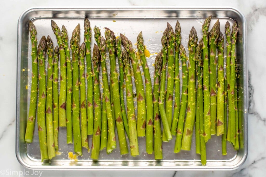 Overhead view of asparagus with oil and seasonings on a baking sheet prepared for Grilled Asparagus recipe.