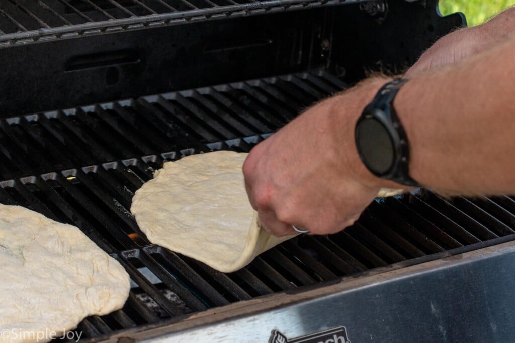 Person's hands putting pizza crust on grill for Grilled Pizza recipe.