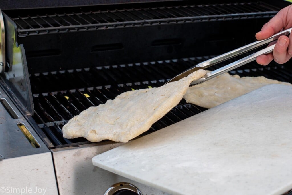 Person's hand using tongs to pull pizza crust from grill for Grilled Pizza recipe.