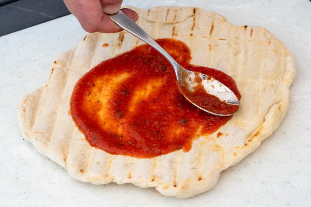 Person's hand using spoon to spread sauce on crust for Grilled Pizza recipe.