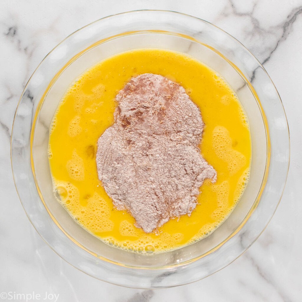 Overhead view of chicken breast with flour on it sitting in bowl of egg wash for Chicken Sandwich recipe.
