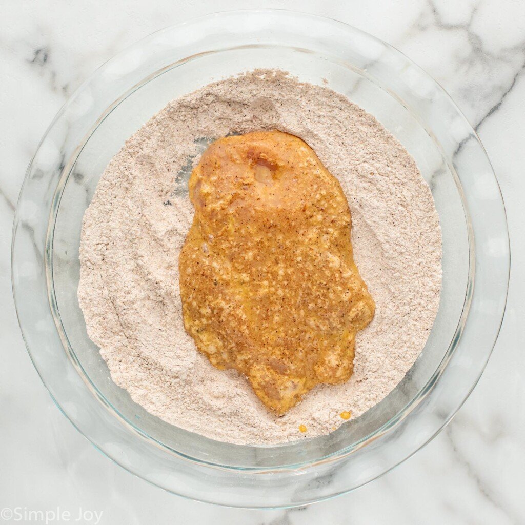 Overhead view of chicken breast with egg wash in bowl of breading for Chicken Sandwich recipe.