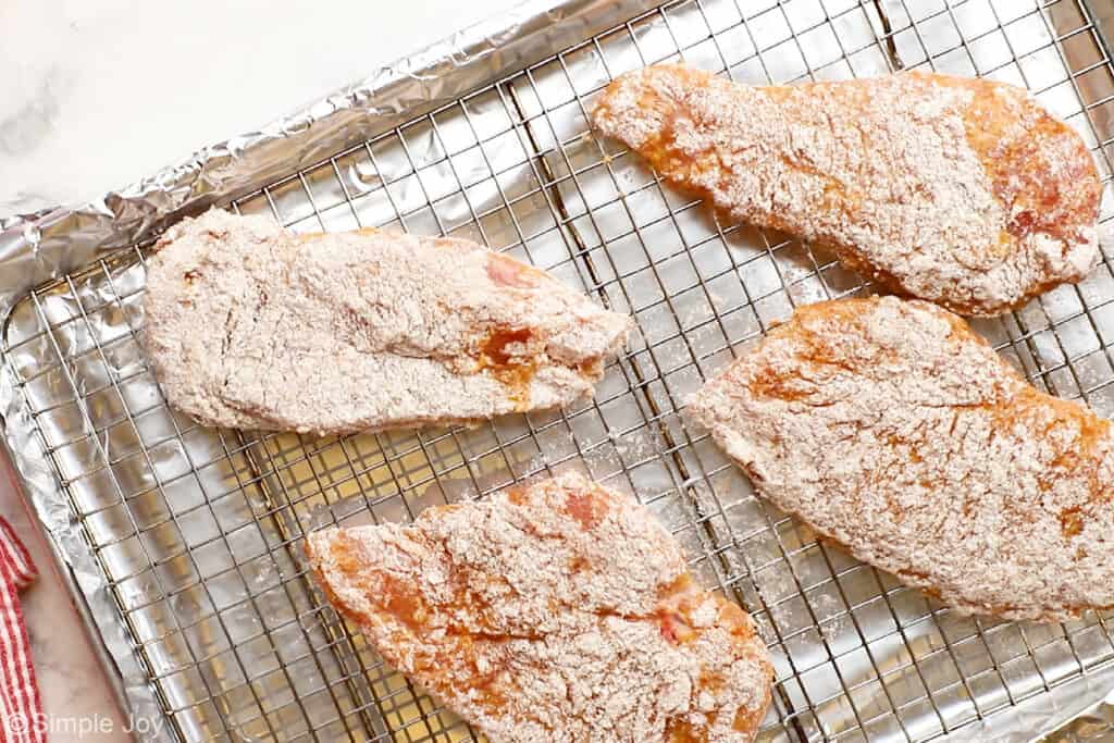 four breaded chicken breasts sitting on a cooling rack inside of a rimmed baking sheet