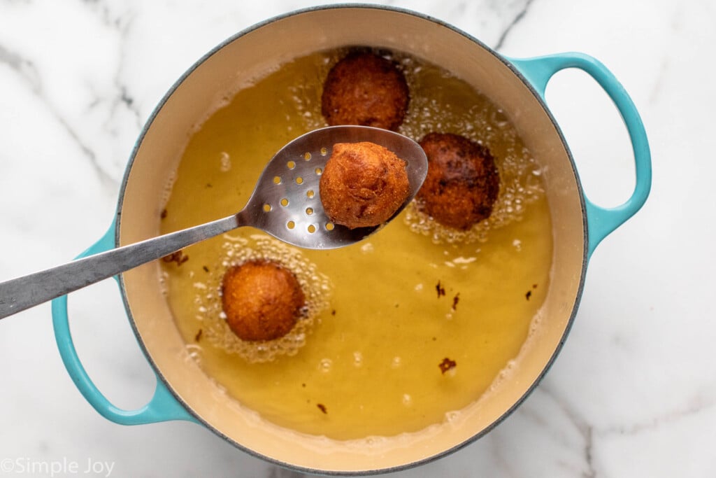 Overhead view of pot of oil with Hush Puppies, with spoon overtop of pot holding one Hush Puppy after cooking.