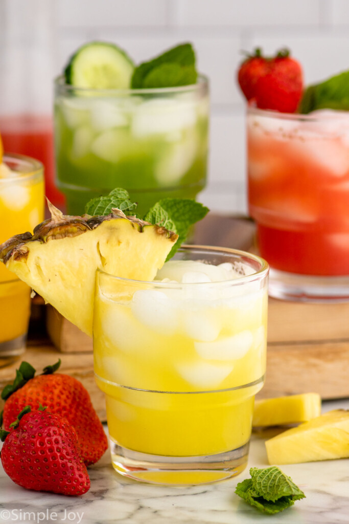 Side view of Agua Fresca drinks garnished with fresh fruit on the rim, with fresh fruit and mint leaves beside as garnish.