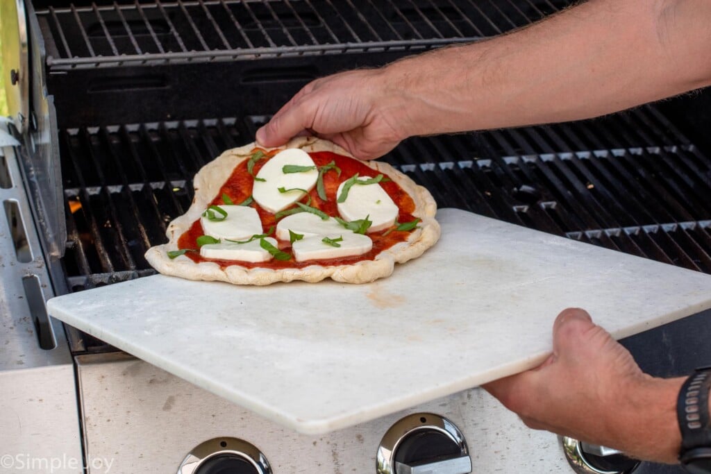 Person sliding pizza with cheese and toppings onto a grill for Grilled Pizza recipe.