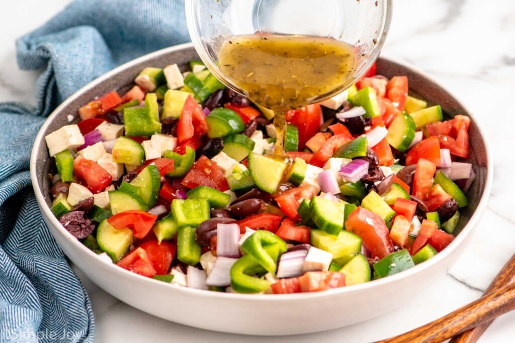 Measuring cup of dressing being poured over chopped vegetables in bowl for Greek Salad recipe.