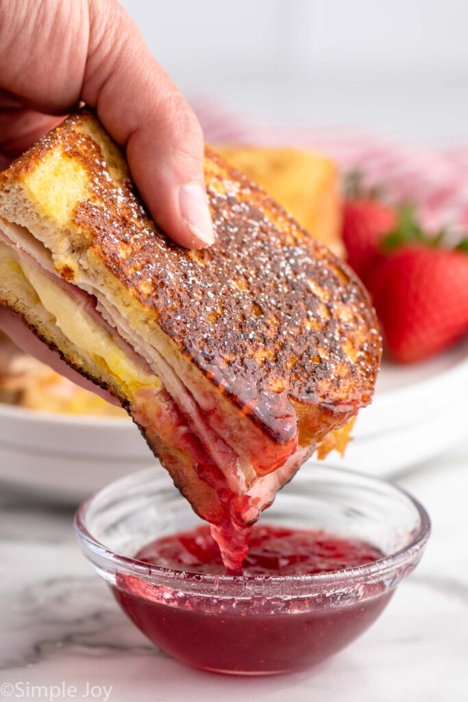 Person's hand dipping half of a Monte Cristo Sandwich into a small dish of sauce.
