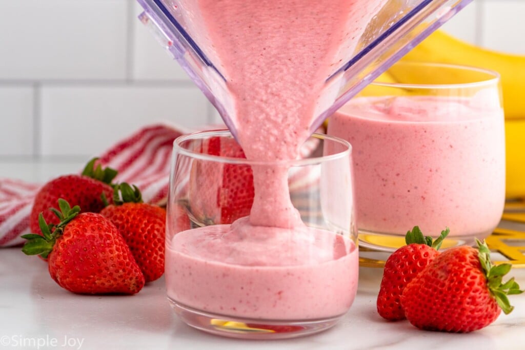 Side view of blender of Strawberry Banana Smoothie recipe being poured into glasses. Strawberries and bananas beside.