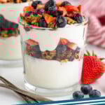 Pinterest graphic for yogurt parfait recipe. Image is side view of yogurt parfait garnished with granola, blueberries, and chopped strawberries with spoons and more berries beside. Text says, "yogurt parfait simplejoy.com."