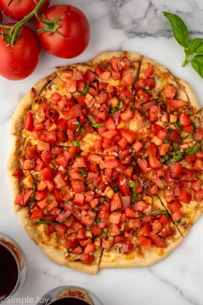 Overhead view of a sliced Bruschetta Pizza. Red wine, tomatoes, and basil leaves beside.