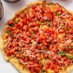 Overhead view of a Bruschetta Pizza that has been cut, garnished with basil and shredded mozzarella cheese. tomatoes, red wine, and basil leaves beside.