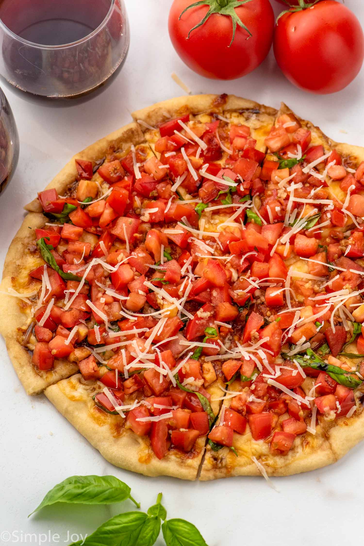 Overhead view of a Bruschetta Pizza that has been cut, garnished with basil and shredded mozzarella cheese. tomatoes, red wine, and basil leaves beside.