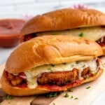 Chicken Parm Sandwiches garnished with parsley flakes on a wooden board with marinara sauce beside