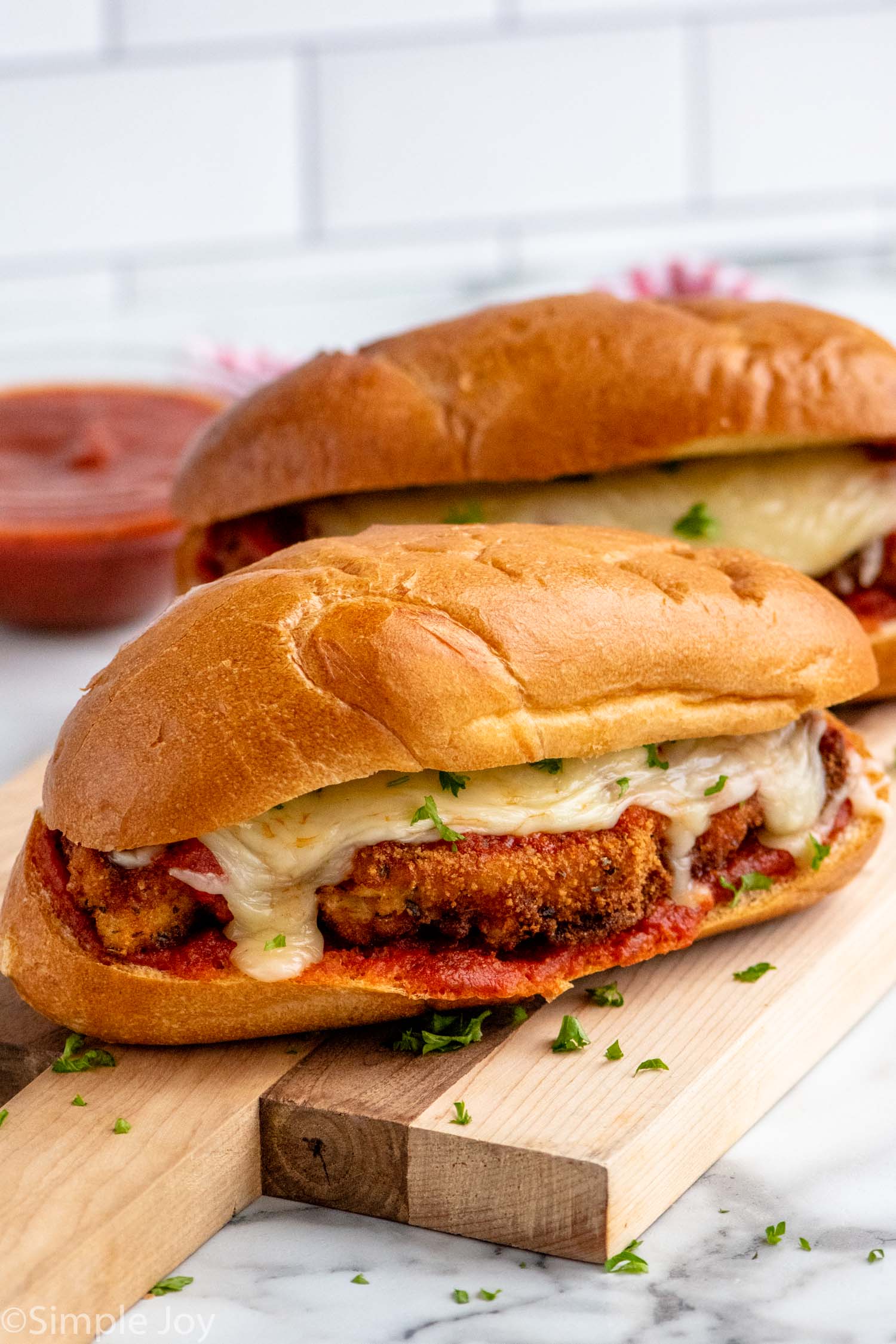 Chicken Parm Sandwiches garnished with parsley flakes on a wooden board with marinara sauce beside