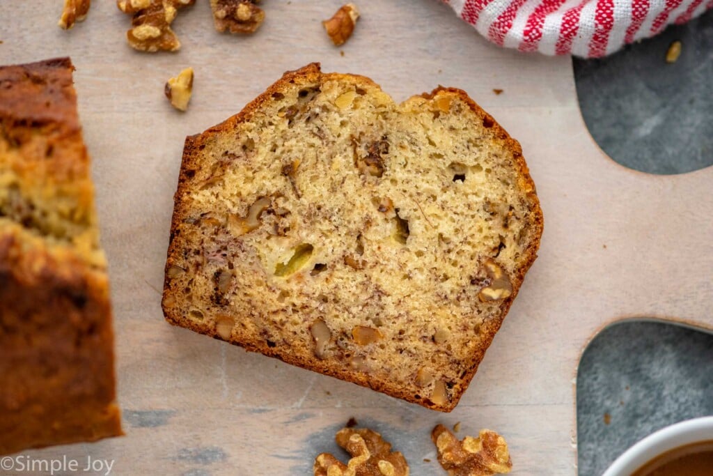 Overhead view of a slice of Banana Nut Bread on a cutting board with a loaf of Banana Nut Bread and walnuts beside.