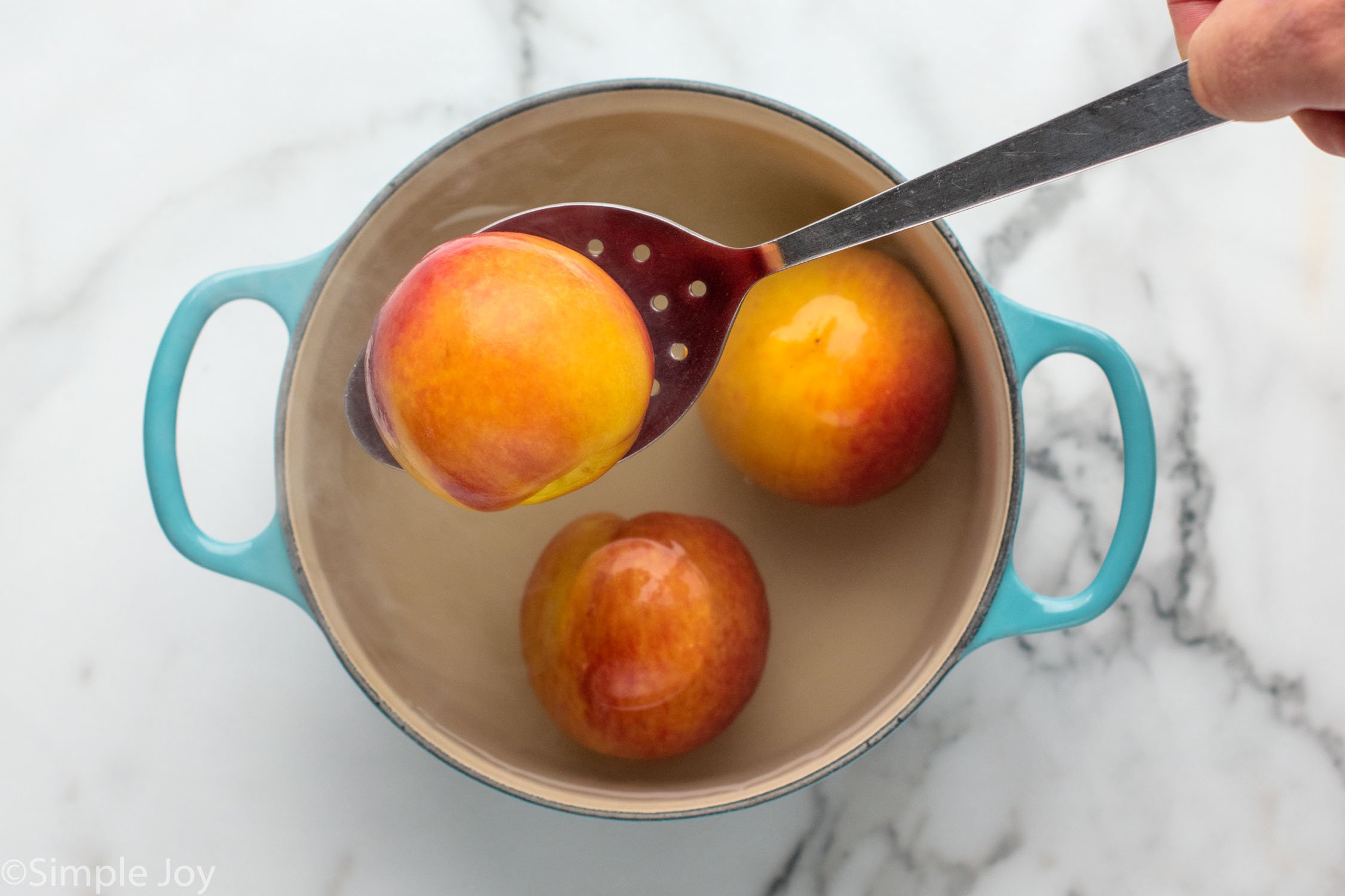 Overhead view of a slotted spoon lifting a peach out of a pot of water and peaches for Peach syrup recipe.