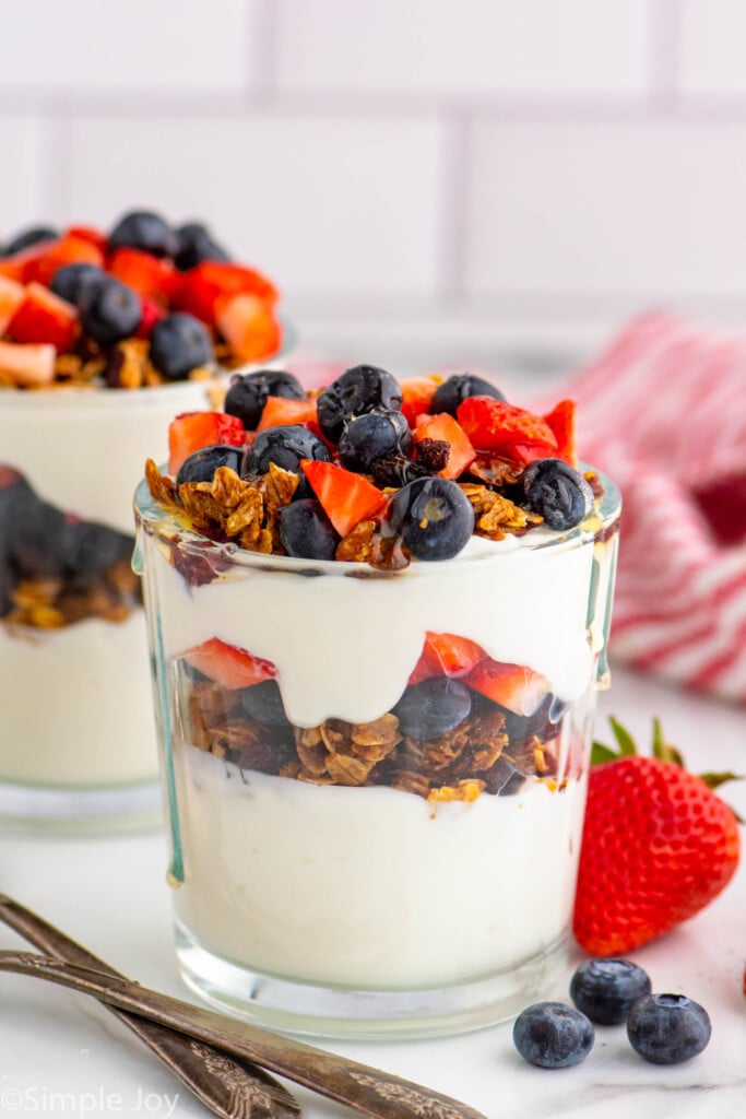 Side view of a yogurt parfait garnished with granola, blueberries, and chopped strawberries. Spoons and more berries beside.