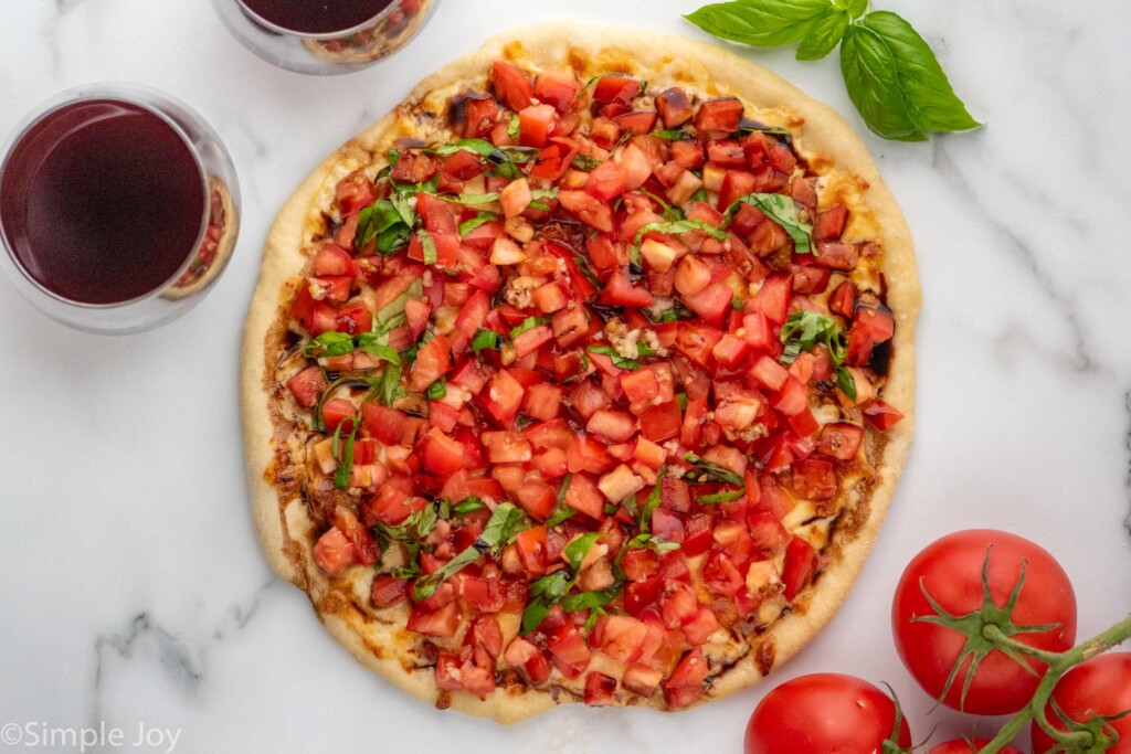 Overhead view of Bruschetta Pizza garnished with basil. Glasses of red wine and tomatoes beside.