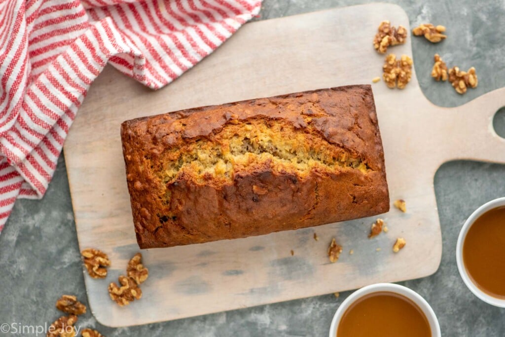 Overhead view of a loaf of Banana Nut Bread on a cutting board with walnuts and two cups of coffee beside.