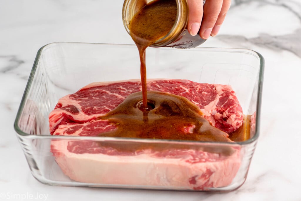Person's hand pouring jar of steak marinade recipe over raw steaks in a glass container.