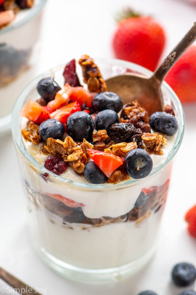 yogurt parfait garnished with chopped strawberries, blueberries, and granola with a spoon. Blueberries and strawberries beside.