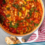 Pinterest graphic for minestrone soup recipe. Image shows overhead view of a pot of minestrone soup with salt and pepper and bread beside. Text says, "absolutely delicious minestrone soup simplejoy.com"
